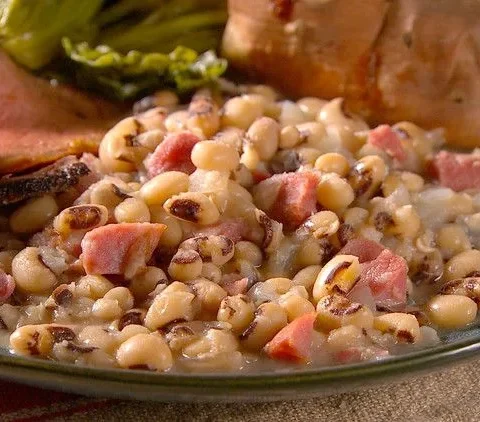 Martha Stewart black-eyed peas with chopped smoked ham hock in a bowl served next to sliced roast beef and a baked sweet potato