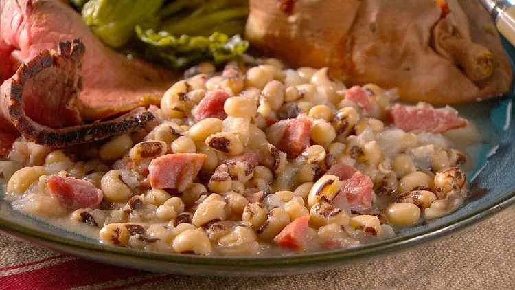 Martha Stewart black-eyed peas with chopped smoked ham hock in a bowl served next to sliced roast beef and a baked sweet potato