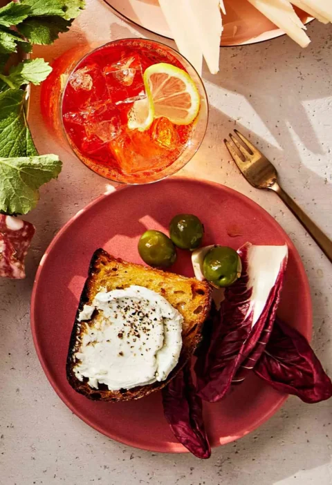 Martha Stewart blue-cheese spread on a toasted slice of bread with black pepper, served on a pink plate with olives and radicchio