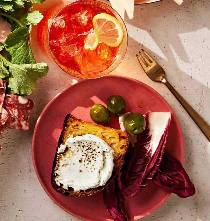 Martha Stewart blue-cheese spread on a toasted slice of bread with black pepper, served on a pink plate with olives and radicchio