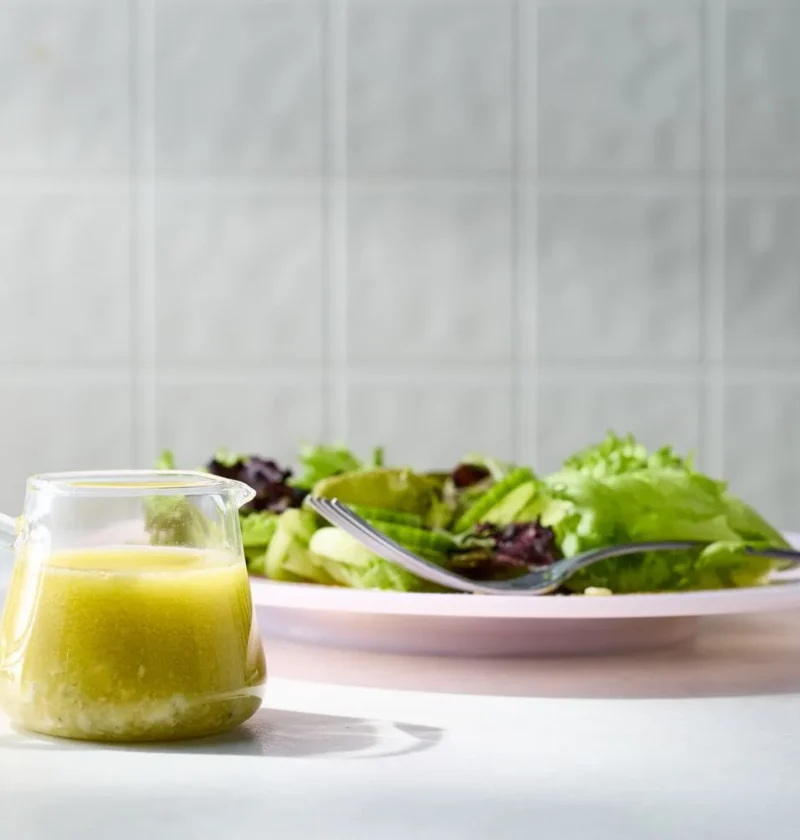 Martha Stewart blue cheese vinaigrette in a small glass pitcher next to a plate of green salad with a fork