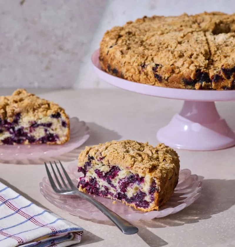 Martha Stewart blueberry buckle with golden streusel topping on a pink cake stand with two slices showing juicy blueberries