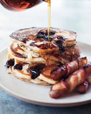 Martha Stewart blueberry buttermilk pancakes stacked on a white plate with maple syrup pouring over the top and bacon on the side