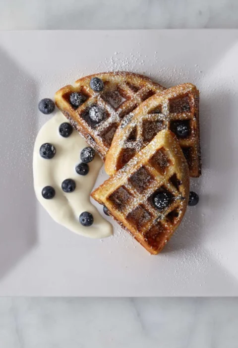 Martha Stewart blueberry buttermilk waffles cut into wedges with fresh blueberries, creme fraiche and powdered sugar on a square white plate