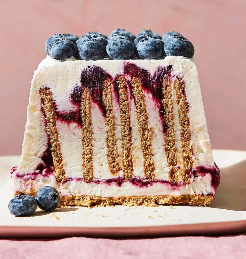 Slice of Martha Stewart blueberry-cheesecake icebox cake showing upright graham crackers, cream cheese layers and blueberry swirls topped with fresh blueberries