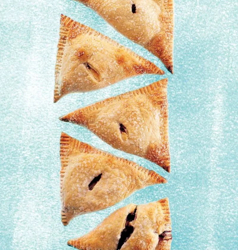 Martha Stewart blueberry-cream cheese hand pies, five golden sugar-topped triangles with vents on a blue background