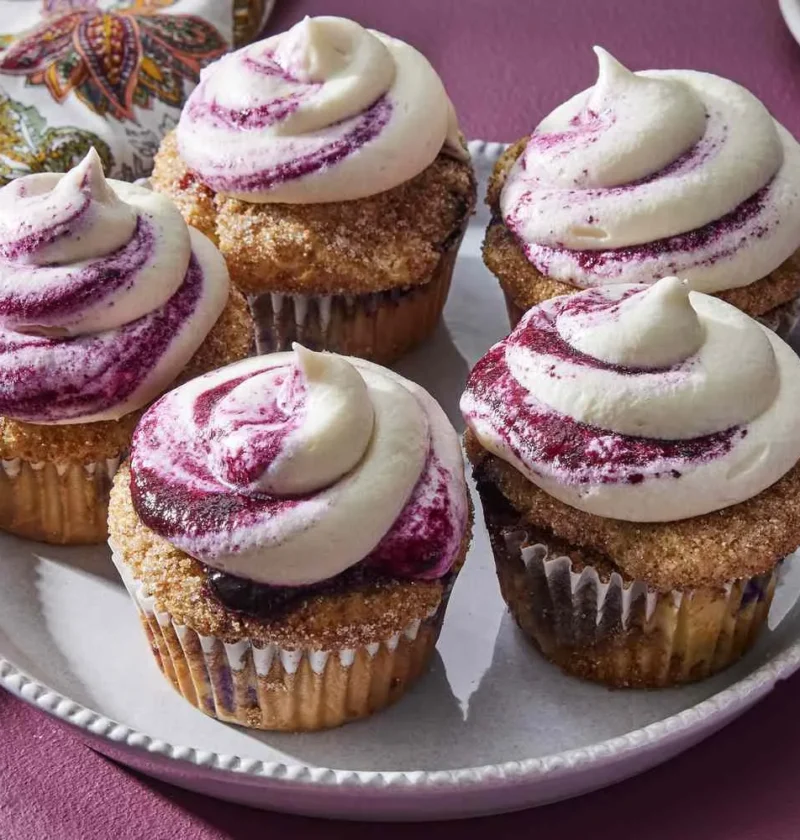 Martha Stewart blueberry cupcakes topped with swirls of cream cheese frosting streaked with blueberry jam on a white plate