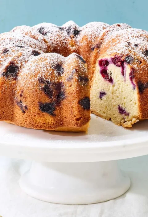 Martha Stewart blueberry-lemon Bundt cake dusted with confectioners' sugar on a white cake stand with one slice cut out