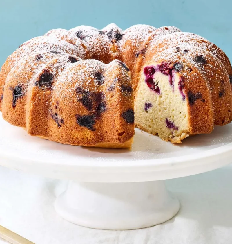 Martha Stewart blueberry-lemon Bundt cake dusted with confectioners' sugar on a white cake stand with one slice cut out