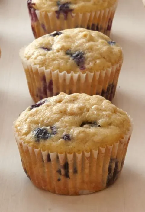 Martha Stewart blueberry-oatmeal muffins in paper liners on a light surface, golden on top with visible blueberries