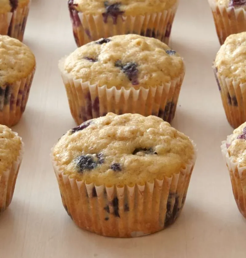 Martha Stewart blueberry-oatmeal muffins in paper liners on a light surface, golden on top with visible blueberries