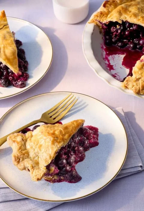 Martha Stewart blueberry pie with a golden double crust, a cut slice showing the dark blueberry filling, and plated slices with gold forks
