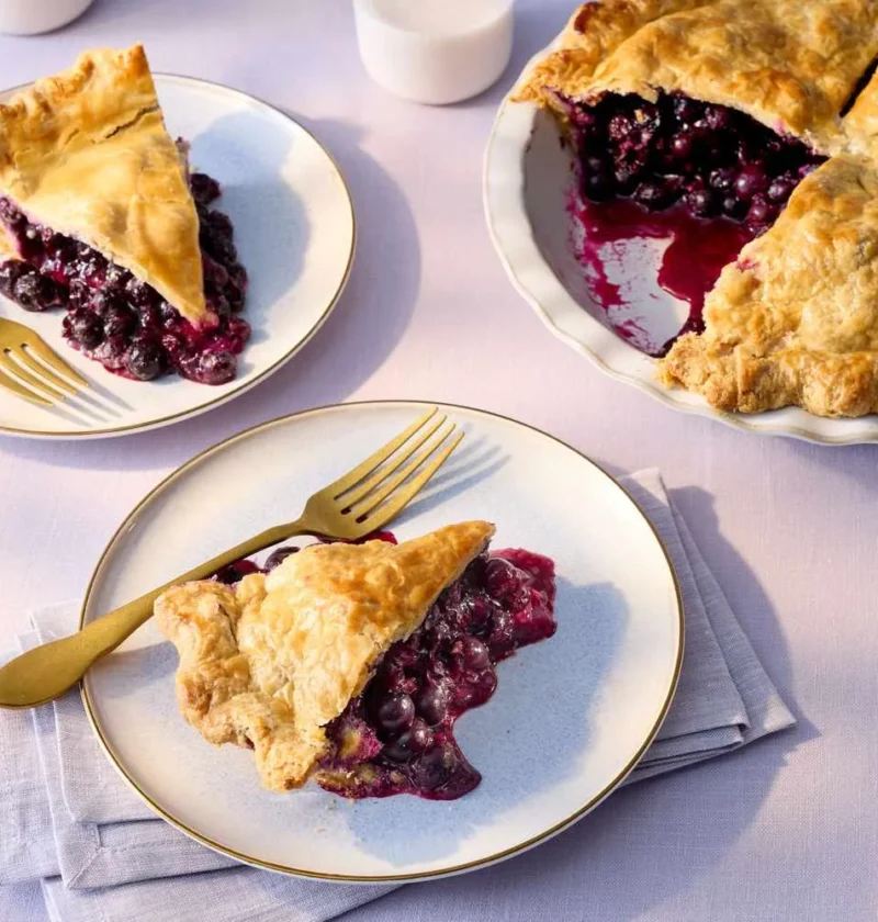 Martha Stewart blueberry pie with a golden double crust, a cut slice showing the dark blueberry filling, and plated slices with gold forks