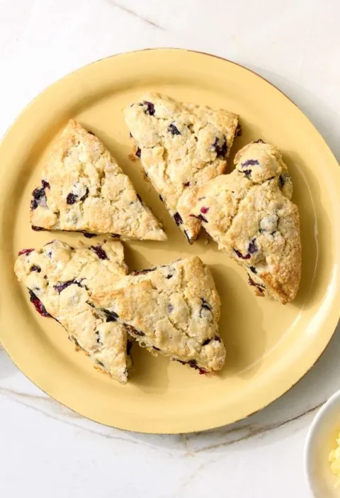 Martha Stewart blueberry scones cut into triangles on a yellow plate with cups of coffee and a dish of softened butter on a marble counter