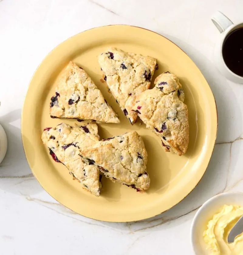 Martha Stewart blueberry scones cut into triangles on a yellow plate with cups of coffee and a dish of softened butter on a marble counter