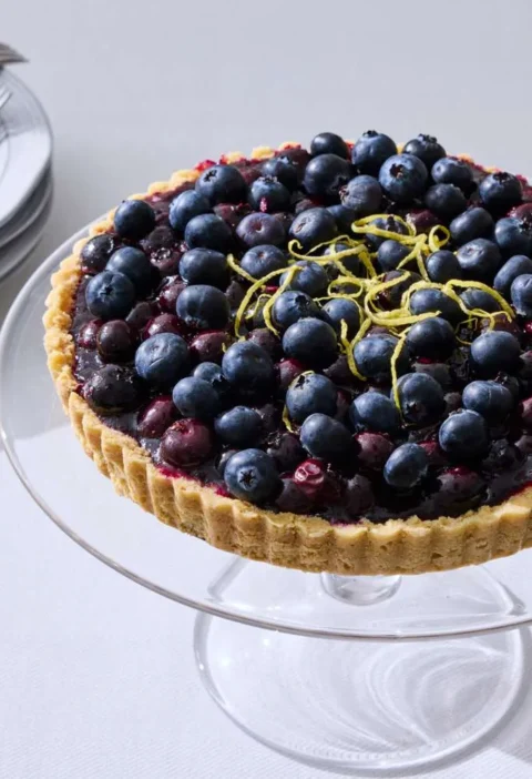 Martha Stewart blueberry tart topped with fresh blueberries and thin lemon zest strips on a glass cake stand
