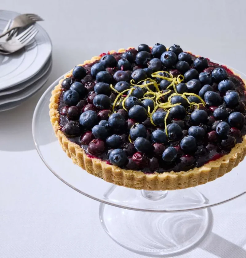 Martha Stewart blueberry tart topped with fresh blueberries and thin lemon zest strips on a glass cake stand