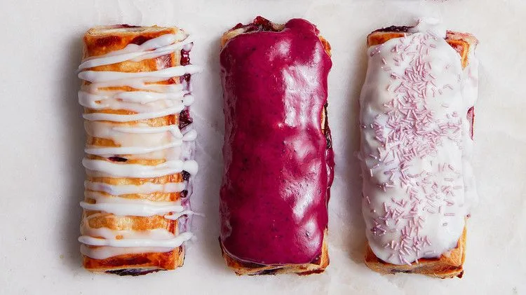 Three Martha Stewart blueberry toaster pastries with white drizzle, purple blueberry glaze and white glaze with purple sprinkles on parchment