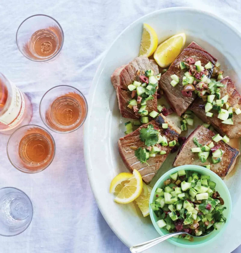 Martha Stewart bluefin tuna with olive, cucumber, and cilantro relish served on an oval platter with lemon wedges next to glasses of rosé