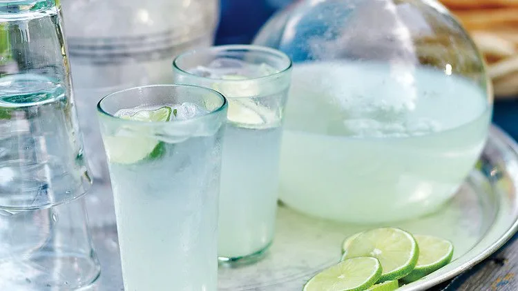 Martha Stewart Bombay Fizz in tall glasses with ice and lime rounds next to a large glass pitcher and sliced limes on a silver tray