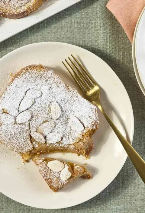 Martha Stewart bostock slice topped with sliced almonds and powdered sugar on a cream plate with gold forks