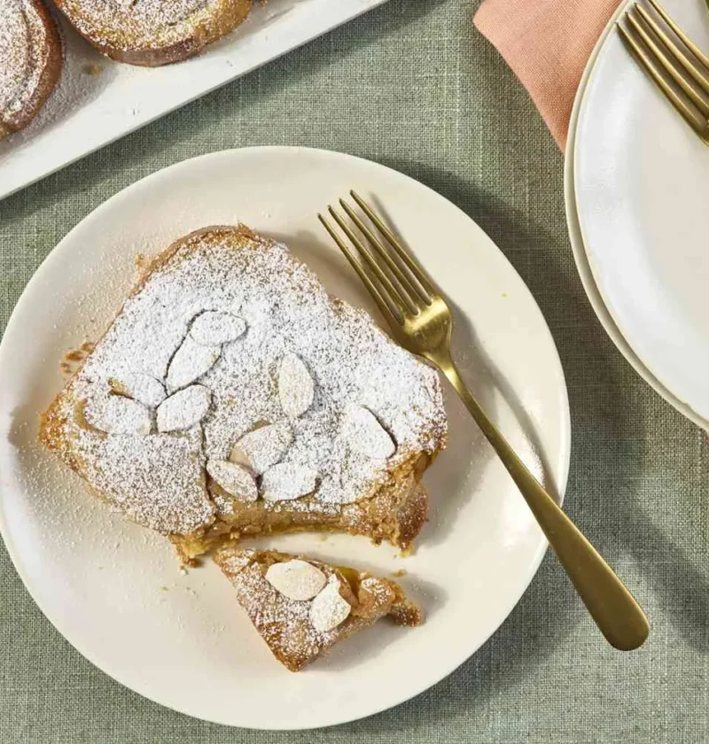 Martha Stewart bostock slice topped with sliced almonds and powdered sugar on a cream plate with gold forks