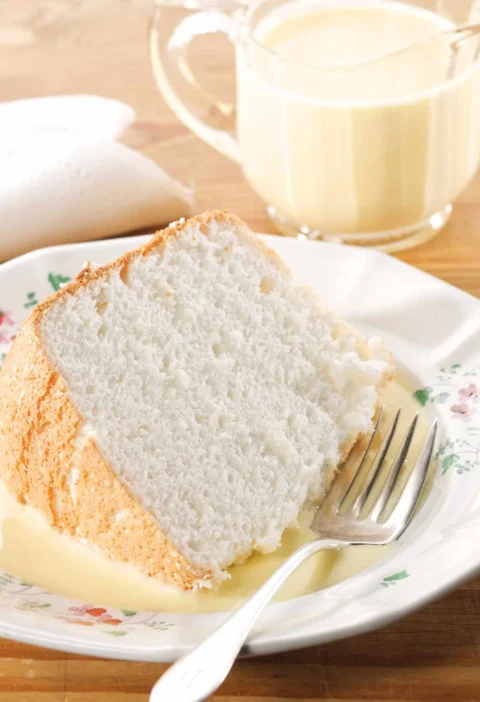 Slice of angel food cake on a floral plate with bourbon creme anglaise pooled around it and a glass pitcher of the sauce behind