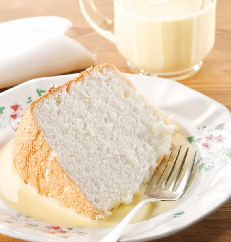 Slice of angel food cake on a floral plate with bourbon creme anglaise pooled around it and a glass pitcher of the sauce behind