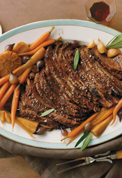 Martha Stewart braised brisket for Passover sliced on an oval platter with roasted baby carrots, parsnips, pearl onions and roasted garlic