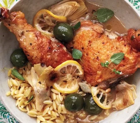 Martha Stewart braised chicken with artichokes, olives and lemon slices served over orzo in a shallow bowl