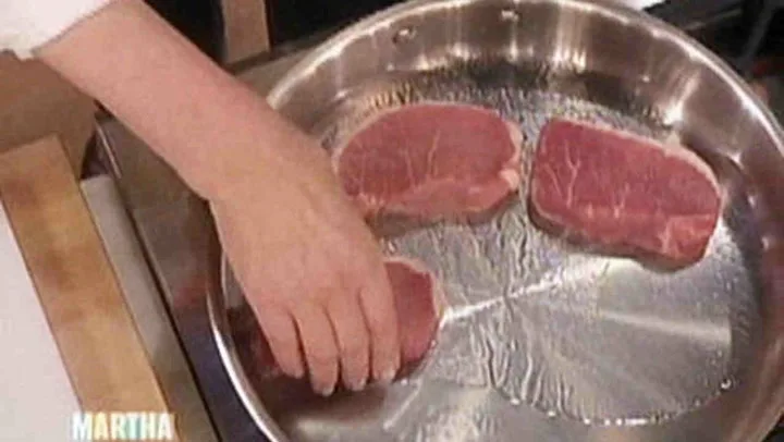 Raw boneless pork chops being placed in a stainless steel skillet for Martha Stewart braised pork chops