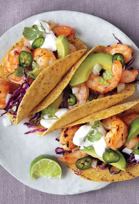 Martha Stewart broiled shrimp tacos in corn tortillas with avocado, red cabbage, sour cream, jalapenos and cilantro on a plate with a lime wedge