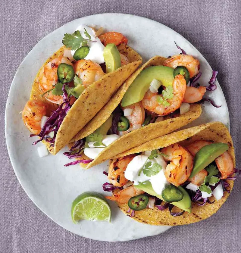 Martha Stewart broiled shrimp tacos in corn tortillas with avocado, red cabbage, sour cream, jalapenos and cilantro on a plate with a lime wedge